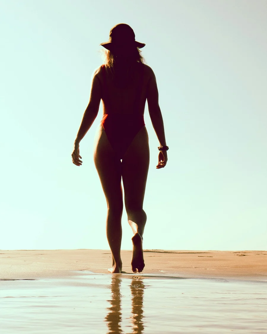 A woman walking toward the light along the shoreline, her reflection in the wet sand.