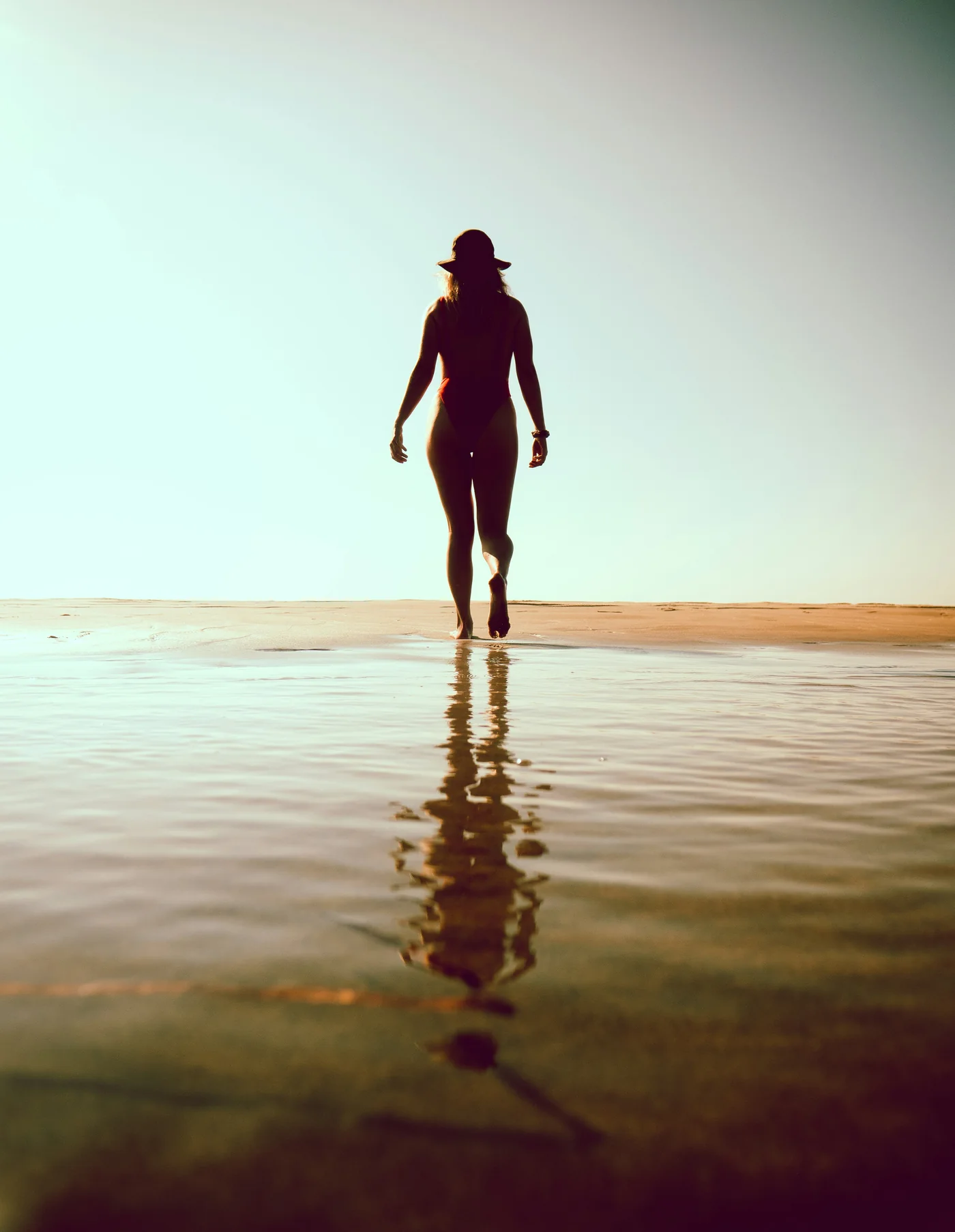 A woman walking toward the light along the shoreline, her reflection in the wet sand.