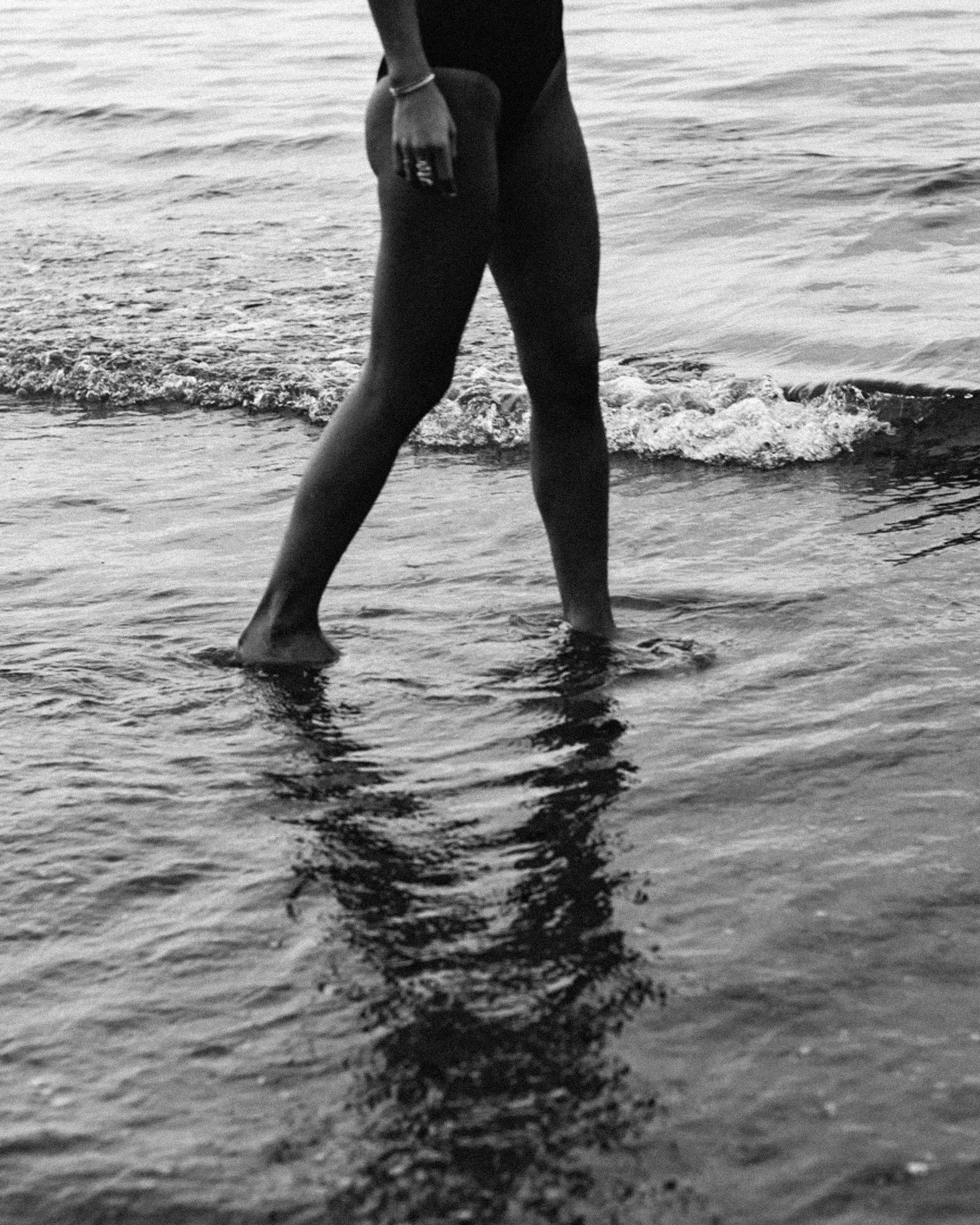 A woman walking through the shallows at the water's edge, her reflection trailing behind her.