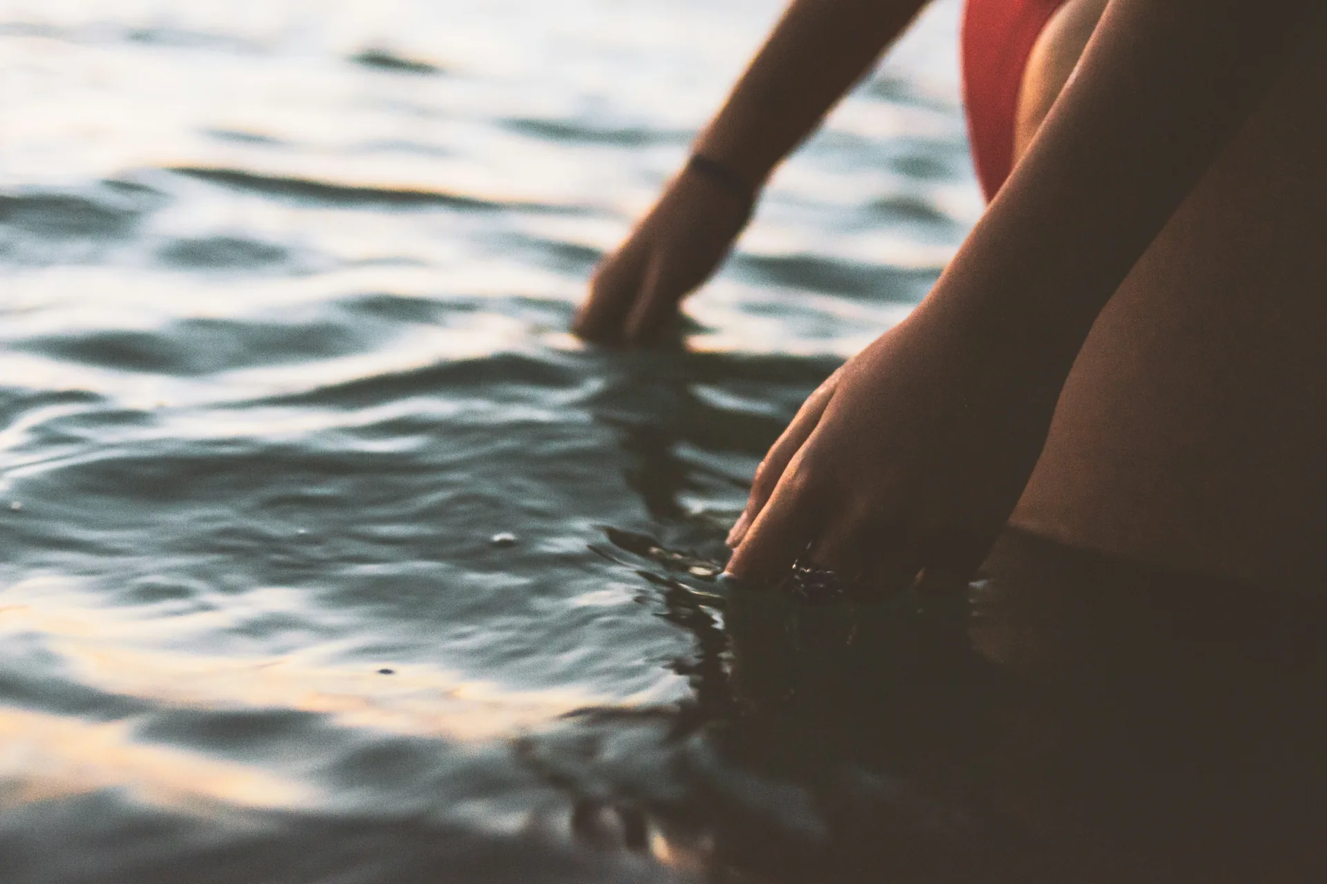 A woman's hands resting on the surface of sunlit water at dusk.