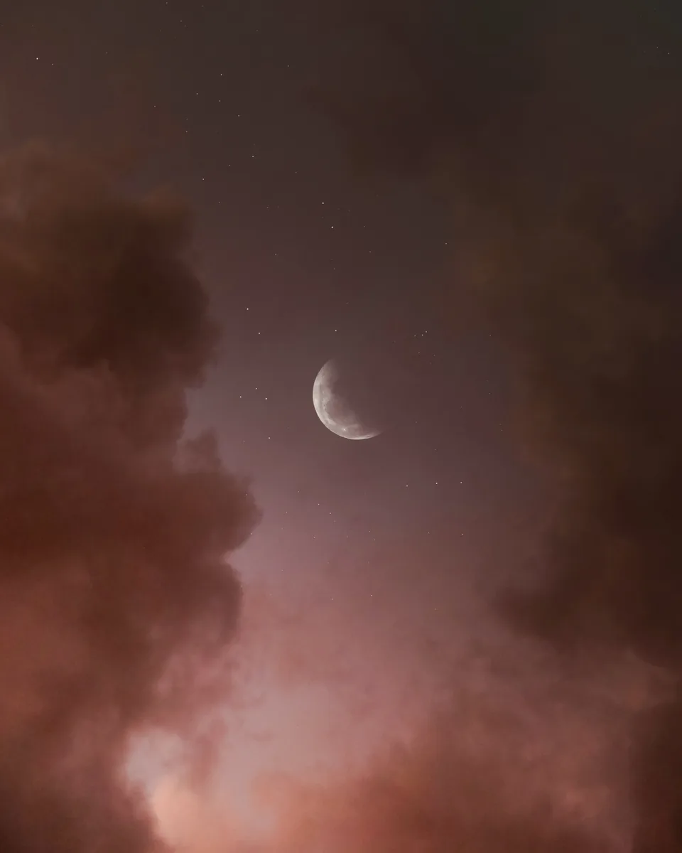 A crescent moon among faint stars, banked by rust-coloured cloud.