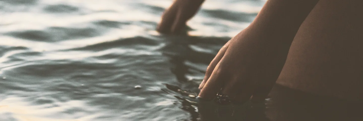 Hands lowered to the surface of the water at dusk.